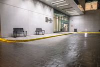 empty benches lined up outside a building at night time on a rainy day with the sidewalk painted yellow