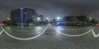 a fish eye lens shows cars turning at an intersection at night, with skyscrapers lit in the background