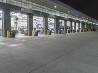 a parking garage with several doors and trash cans around it at night time as traffic passes by