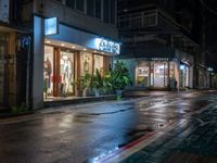 a wet street with store fronts and lights on, at night, in the rain