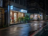 a wet street with store fronts and lights on, at night, in the rain