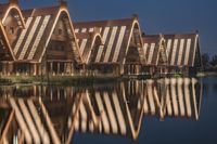 a group of houses on stilts near water at night time with lights reflected in the glass