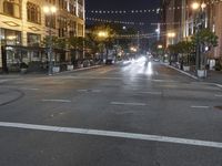 an intersection with many lights hanging on the buildings at night, with people standing on the sidewalk