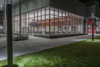 a building with several glass windows next to some grass and trees at night time on a field in front of a street lamp