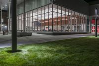 a building with several glass windows next to some grass and trees at night time on a field in front of a street lamp