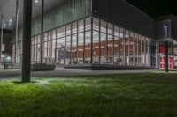 a building with several glass windows next to some grass and trees at night time on a field in front of a street lamp