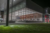 a building with several glass windows next to some grass and trees at night time on a field in front of a street lamp