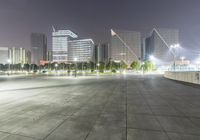 a person sitting on a skate board in an empty parking lot at night time with street lights and buildings around