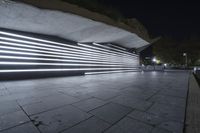 a large walkway in front of a building at night with neon light and concrete on the sidewalk