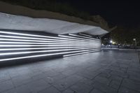 a large walkway in front of a building at night with neon light and concrete on the sidewalk