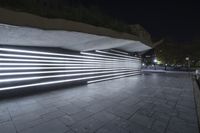 a large walkway in front of a building at night with neon light and concrete on the sidewalk