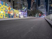 an empty roadway at night with lights shining on buildings in the background and a colorful mural