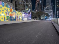 an empty roadway at night with lights shining on buildings in the background and a colorful mural