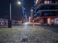 the sidewalk has several bricks on it at night near many tall buildings and a clock tower