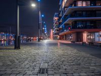 a long brick walkway with lights on, in an urban setting at night by the waterfront