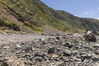 a rock strewn trail leads to a rocky cliff slope in the mountains above is a body of water and green cliffs