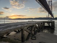 a wooden dock under a bridge over water with boats docked at sunset below it and sun set