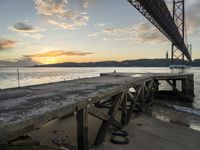 a wooden dock under a bridge over water with boats docked at sunset below it and sun set