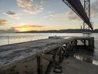 a wooden dock under a bridge over water with boats docked at sunset below it and sun set