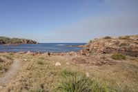 the coast of an ocean surrounded by rocks and grass with an airplane in the distance