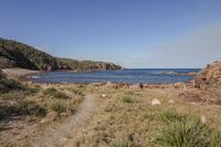 the coast of an ocean surrounded by rocks and grass with an airplane in the distance