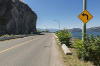 the sign points at a bend on the side of a road near the ocean and mountains