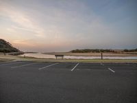 an empty parking lot next to the ocean at sunset with no cars around it and several picnic benches near it