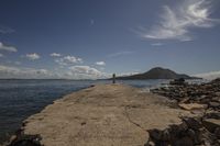 a lone pier at the ocean in front of mountains and water and clouds above it