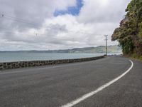 a couple people are walking down a paved road by the ocean, as a motorcycle passes