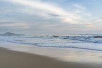 an ocean view of a sandy beach, waves lapping on the shore and distant clouds in the sky