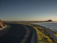 a long winding road next to the ocean at sunset time, next to an entrance for a beach