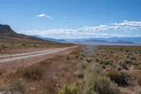 a dirt road leading through an open desert area, with mountains in the background and blue skies above