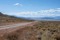 a dirt road leading through an open desert area, with mountains in the background and blue skies above