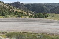 a small empty dirt field and a forest behind it on a sunny day with hills and mountains in the background