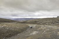 the man is riding the motorcycle in the desert under the cloudy sky and hills with rocks and gravel