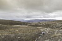 the man is riding the motorcycle in the desert under the cloudy sky and hills with rocks and gravel