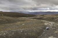 the man is riding the motorcycle in the desert under the cloudy sky and hills with rocks and gravel