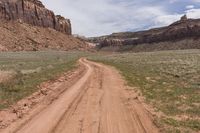 dirt road next to red canyon with green grass and a sign on it that reads