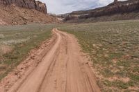 dirt road next to red canyon with green grass and a sign on it that reads