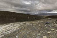 a dirt road through a barren terrain area with rocks and grass around it and two mountains in the distance