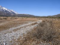 a mountain range with grass and dry bushes with a trail leading to it and snow capped mountains beyond