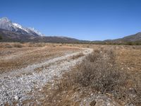 a mountain range with grass and dry bushes with a trail leading to it and snow capped mountains beyond