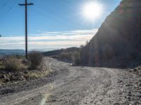 a narrow gravel road with rocks, grass and telephone pole in the background and mountains