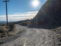 a narrow gravel road with rocks, grass and telephone pole in the background and mountains