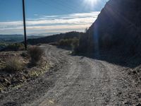 a narrow gravel road with rocks, grass and telephone pole in the background and mountains