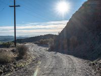 a narrow gravel road with rocks, grass and telephone pole in the background and mountains