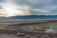 the sun sets behind mountains in a desert landscape with green grass and scrubby weeds