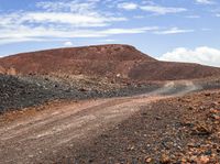 a dirt road surrounded by rocks and red mountains with clouds in the background under a partly cloudy sky