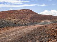 a dirt road surrounded by rocks and red mountains with clouds in the background under a partly cloudy sky