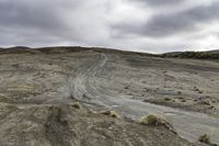 two motorcycles driving on dirt and sand near rocks and a hill and shrubs under cloudy skies